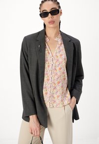 Grå rutete blazer med strukturert passform, kombinert med en lys blomstret skjorte med knapper. Beige bukser og svarte rektangulære solbriller fullfører antrekket.