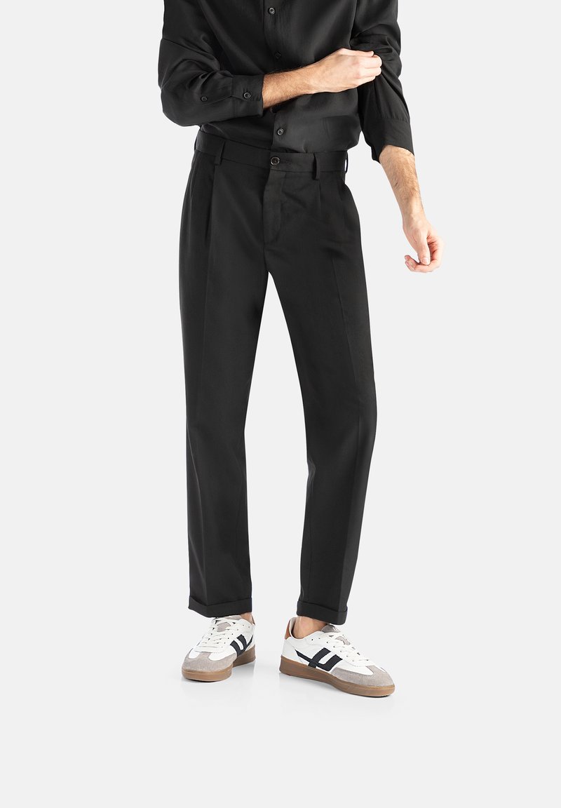 Černé kalhoty na míru s rovnými nohavicemi, dvojitými záhyby a hladkým povrchem, v kombinaci s bílými a šedými teniskami.