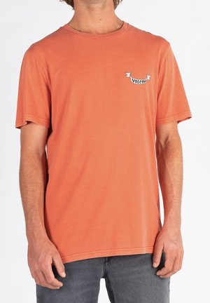 Mand iført en enkel orange Volcom t-shirt og grå bukser mod en hvid baggrund, hænder afslappet ved hans sider.