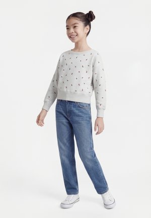 Fille souriante avec des cheveux en chignons, portant un sweat léger avec de petites cerises rouges, un jean bleu et des baskets blanches, debout sur un fond blanc.