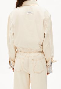 Beige denimjacka med elastisk midja, har en logotyp på baksidan och ljusblå rutmönstrade manschetter. Strukturerad yta, utan synlig hårdvara.