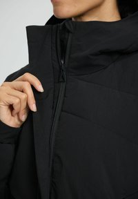 Die Hand einer Person hält den aufgezogenen Kragen einer schwarzen Daunenjacke und zeigt den Reißverschluss sowie das hochgeschlossene Design.