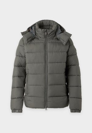Jeune homme debout portant une doudoune gris foncé, un t-shirt blanc, un jean bleu ample et des baskets grises sur un fond clair uni.