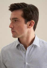 Witte shirt met blauwe ronde patronen, kraag en knoopsluiting aan de voorkant, geweven stof structuur, zijprofiel van een man.