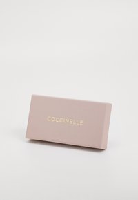 Rosa rechteckige Box mit matter Oberfläche, die den Markennamen "COCCINELLE" in goldener Schrift zeigt. Einfaches und sauberes Design ohne zusätzliche Details.