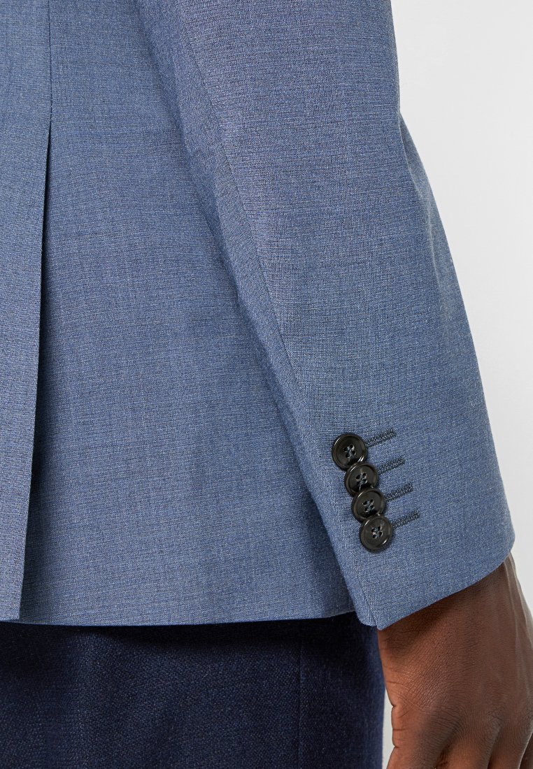 Ärmel eines blauen Blazers mit vier schwarzen Knöpfen in der Nähe des Bündchens und einer daneben ruhenden Hand, kontrastiert mit dunklen Hosen.