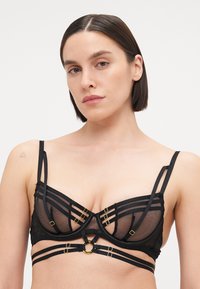 Soutien-gorge noir en matière résille avec armatures structurées, plusieurs designs de bretelles, accents en métal doré et connecteur circulaire.