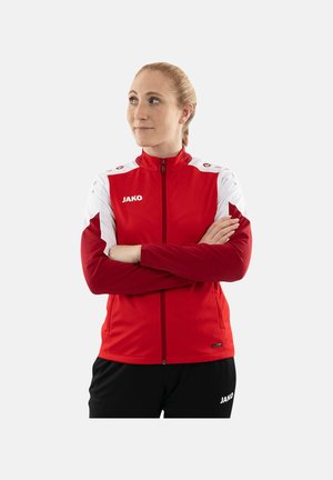 Femme aux cheveux blonds attachés en queue de cheval, portant une veste de sport zippée rouge et blanche et un pantalon noir, debout les bras croisés, regardant légèrement à gauche.