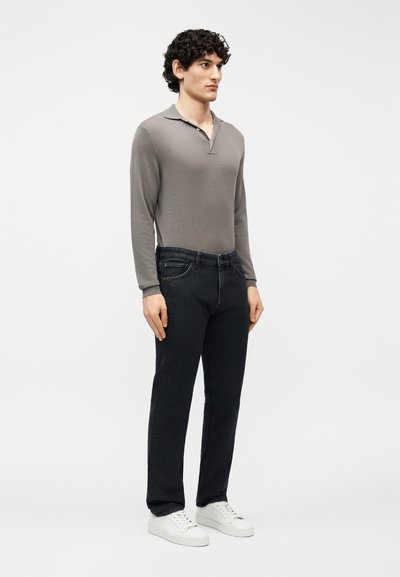 Νεαρός άνδρας που στέκεται, φοράει γκρι μακρυμάνικο πόλο πουκάμισο, μαύρα τζιν και λευκά αθλητικά παπούτσια μπροστά σε απλό λευκό φόντο.