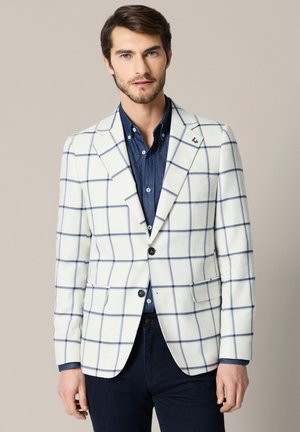 Mężczyzna w białej marynarce z granatowym wzorem w kratę na niebieskiej koszuli i ciemnych spodniach, stojący na jednolitym jasnym tle.