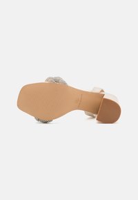 Lys beige sandaler med en glatt overdel, dekorert med en perlesmykke rundt ankelen, polstret såle og teksturert gummisåle.