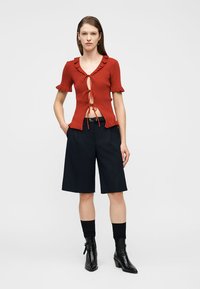 Mujer con blusa roja de manga corta con lazos delanteros, shorts negros hasta la rodilla, calcetas negras y botines negros, de pie sobre un fondo liso.