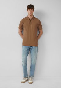 Braunes Poloshirt mit einem Reißverschlusskragen, kombiniert mit hellblauen Jeans und beigen Sneakers. Das Model steht vor einem schlichten weißen Hintergrund.