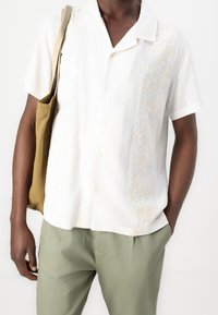 Biała koszula z krótkim rękawem z misternym beżowym kwiatowym haftem, zestawiona z oliwkowozielonymi spodniami i brązową torebką na ramię.