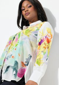Camicia leggera e trasparente con stampa floreale in colori vivaci che includono giallo, rosa e verde; presenta un colletto e polsini bianchi.