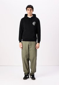 Felpa nera con cappuccio e coulisse, dotata di una tasca frontale, con un logo bianco. Abbinata a pantaloni larghi di colore verde oliva e sneakers nere.