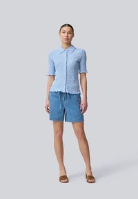 Camisa de botão texturizada azul clara com mangas curtas, combinada com calções de ganga claros e sandálias planas castanhas. Conjunto simples e casual.