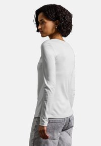 Vit långärmad skjorta, mjukt bomullstyg, rund halsringning, slim fit, kombinerad med ljusgrå jeans och en strukturerad lockig frisyr.
