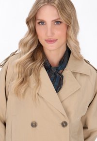 Beige dobbeltbrystet trenchcoat med store knapper, skulderepauleter og et mørkt ternet tørklæde bundet om halsen. Glat stoffinish.