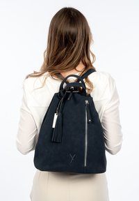 En svart ryggsäck i konstläder med texturerad yta, runda handtag, justerbara remmar och en sidoficka med dragkedja prydd med en tofsdetalj.