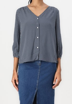 Kvinde iført en marineblå mønstret skjortebluse med trekvartlange ærmer og en højtaljet blå denimnederdel med frontslids.