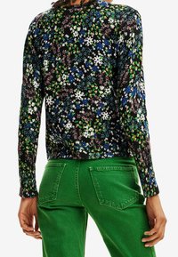 Kobieta ubrana w czarną bluzkę z długim rękawem z wielokolorowym kwiatowym wzorem oraz jasne zielone spodnie, widziana od tyłu.