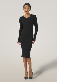 Abito midi a coste nero con maniche lunghe, scollo a barca e silhouette aderente, abbinato a tacchi alti a punta.