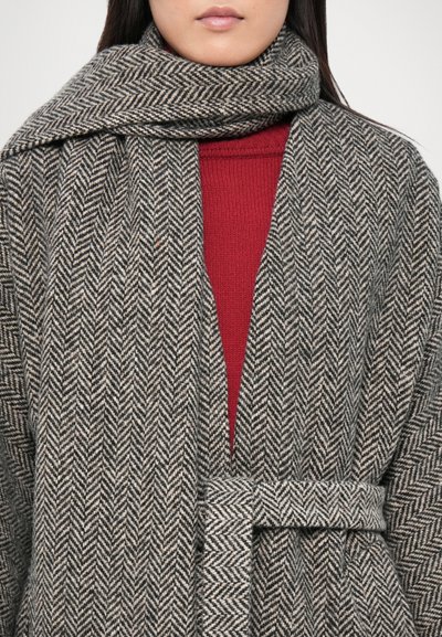 Manteau en laine avec un motif en chevrons gris et crème, présentant un pull rouge en dessous et une ceinture assortie pour le fermer.