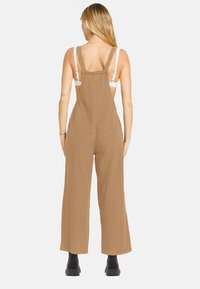 Brown cotton jumpsuit with wide legs, adjustable straps, and a relaxed fit, featuring a backless design and textured fabric.