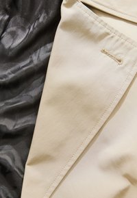 Una tela beige con una textura suave presenta un borde cosido visible y un pequeño detalle de lazo; junto a ella hay un material negro, con patrón, transparente.