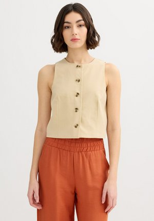 Mujer de cabello corto y oscuro que lleva una blusa sin mangas beige con botones y pantalones de talle alto color óxido, de pie contra un fondo liso.