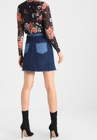 Jupe en denim bleu foncé avec un ourlet brut et deux poches arrière ; portée avec un top transparent à manches longues à motifs floraux et des bottes à talons hauts noires.