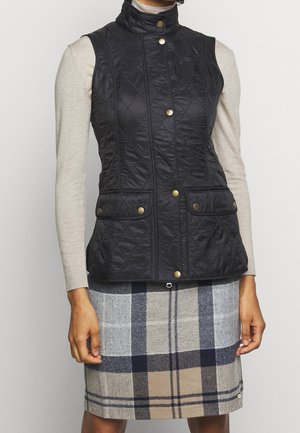 Woman wearing a black quilted sleeveless vest with gold buttons over a long-sleeve beige top, paired with a beige and navy plaid skirt.