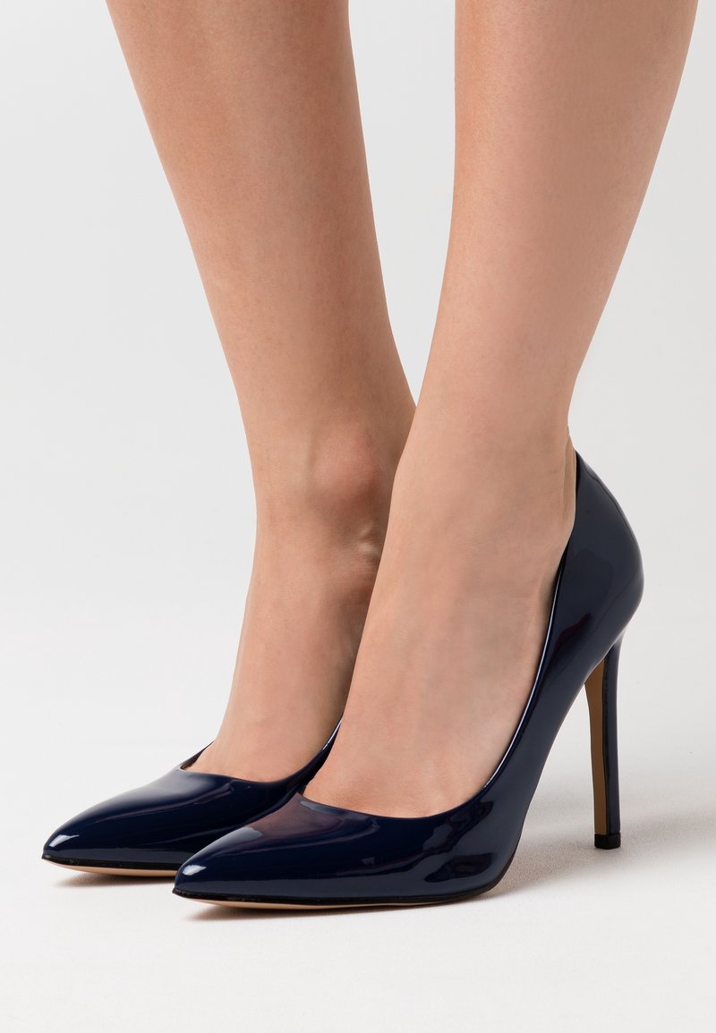 Pieds d'une femme portant des escarpins brillants bleu marine à bout pointu avec talons aiguilles sur un fond clair.