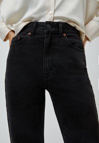 Jeans de mezclilla negra con una cintura alta, diseño de cinco bolsillos y adornos de botones de cobre, exhibidos con una camisa de color claro.