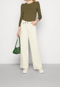 Frau in einem olivgrünen Langarmoberteil und cremefarbenen, weiten Hosen, die eine kleine grüne Handtasche hält und weiße Plattformschuhe trägt.