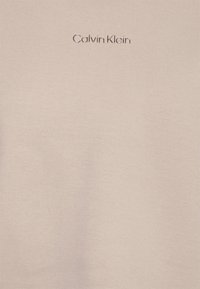Camiseta de algodón beige con una textura suave, que presenta un sutil logo de Calvin Klein en un color de hilo a juego en el lado superior izquierdo.