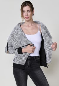 Szürke és fehér mintás dzseki nyitott elülső résszel, fekete szegéllyel és bordás mandzsettával. Fehér felsőre és sötét farmert visel.