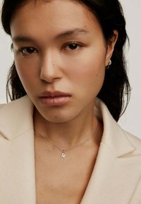 Collar de plata con un colgante en forma de letra "M", llevado sobre un abrigo beige claro. La modelo tiene el cabello largo y oscuro.