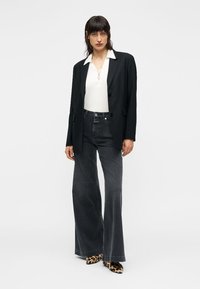 Μαύρο σακάκι πάνω από λευκή μπλούζα, συνδυασμένο με γκρι παντελόνι με φαρδιά πόδια και παπούτσια με λεοπάρ μοτίβο. Λείες υφές και αντιθετικά χρώματα.