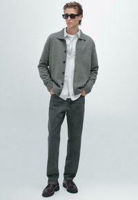 Giovane uomo con occhiali da sole neri, cardigan grigio con bottoni, camicia a righe bianche, jeans grigio scuro e scarpe nere, in piedi con le mani nelle tasche.