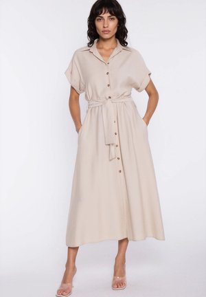 Kobieta w beżowej, midi sukience zapinanej na guziki, z krótkimi rękawami, zawiązanym paskiem w talii i przezroczystymi sandałami na wysokim obcasie, stojąca z rękami w kieszeniach.