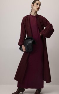 Longue robe bordeaux avec manches bouffantes à couches, assortie d'un long manteau assorti. Sac structuré noir et bottes marron montantes au genou complètent le look.