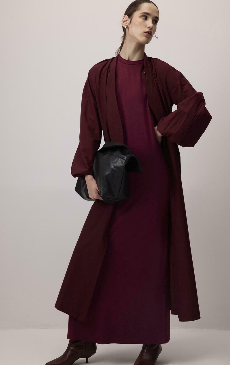 Longue robe bordeaux avec manches bouffantes à couches, assortie d'un long manteau assorti. Sac structuré noir et bottes marron montantes au genou complètent le look.