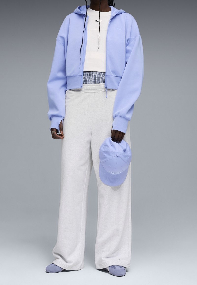 Personne portant un sweat à capuche bleu clair zippé, un crop top blanc, un pantalon large gris clair, tenant une casquette bleue et des chaussures bleues assorties, sur fond gris.
