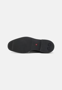 Semelle de chaussure en caoutchouc noir avec surface texturée, forme arrondie et accent rouge près du talon. Marque visible sur le côté.