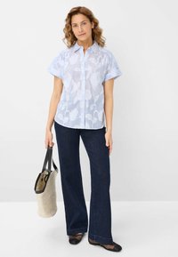 Lichtblauw overhemd met bloempatroon, korte mouwen en knoopsluiting, gecombineerd met donkere spijkerbroek met flares en een beige tote bag.