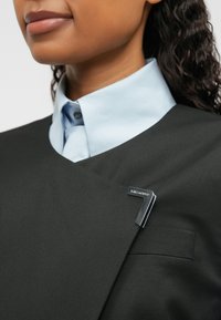 Čierny strihaný odev s ostrým V-výstrihom, vrstvený cez svetlomodrú košeľu. Výrazná strieborná brošňa v tvare čísla sedem na ľavej strane.