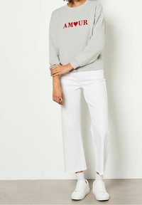 Personne portant un sweat-shirt gris clair avec "AMOUR" et un cœur rouge, un pantalon blanc court, des chaussettes blanches et des baskets blanches, debout à l'intérieur.