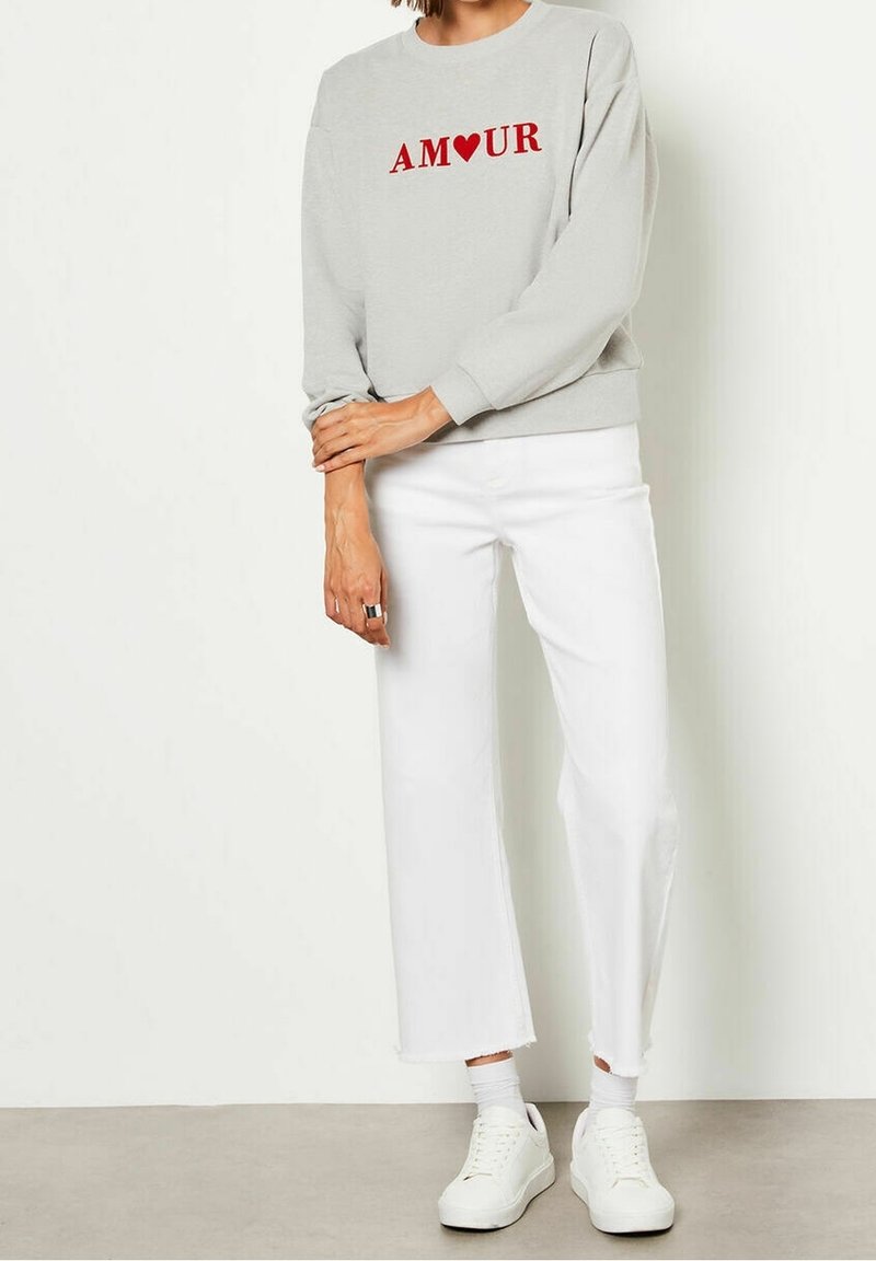 Personne portant un sweat-shirt gris clair avec "AMOUR" et un cœur rouge, un pantalon blanc court, des chaussettes blanches et des baskets blanches, debout à l'intérieur.